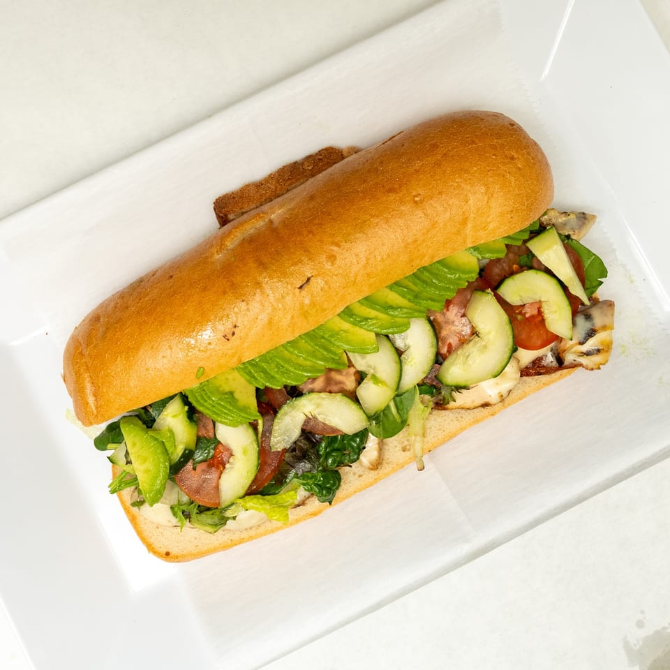 A large submarine sandwich filled with avocado, cucumber, lettuce, tomato, and meat on a white plate.