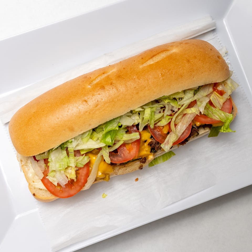A long submarine sandwich filled with meat, melted cheese, shredded lettuce, and sliced tomatoes on a white tray.